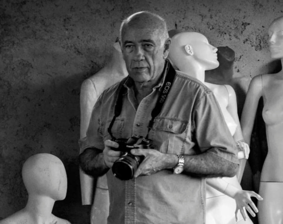 Alexis Pérez-Luna, con camisa clara y una cámara en las manos, posa entre maniquíes blancos en una foto en blanco y negro.