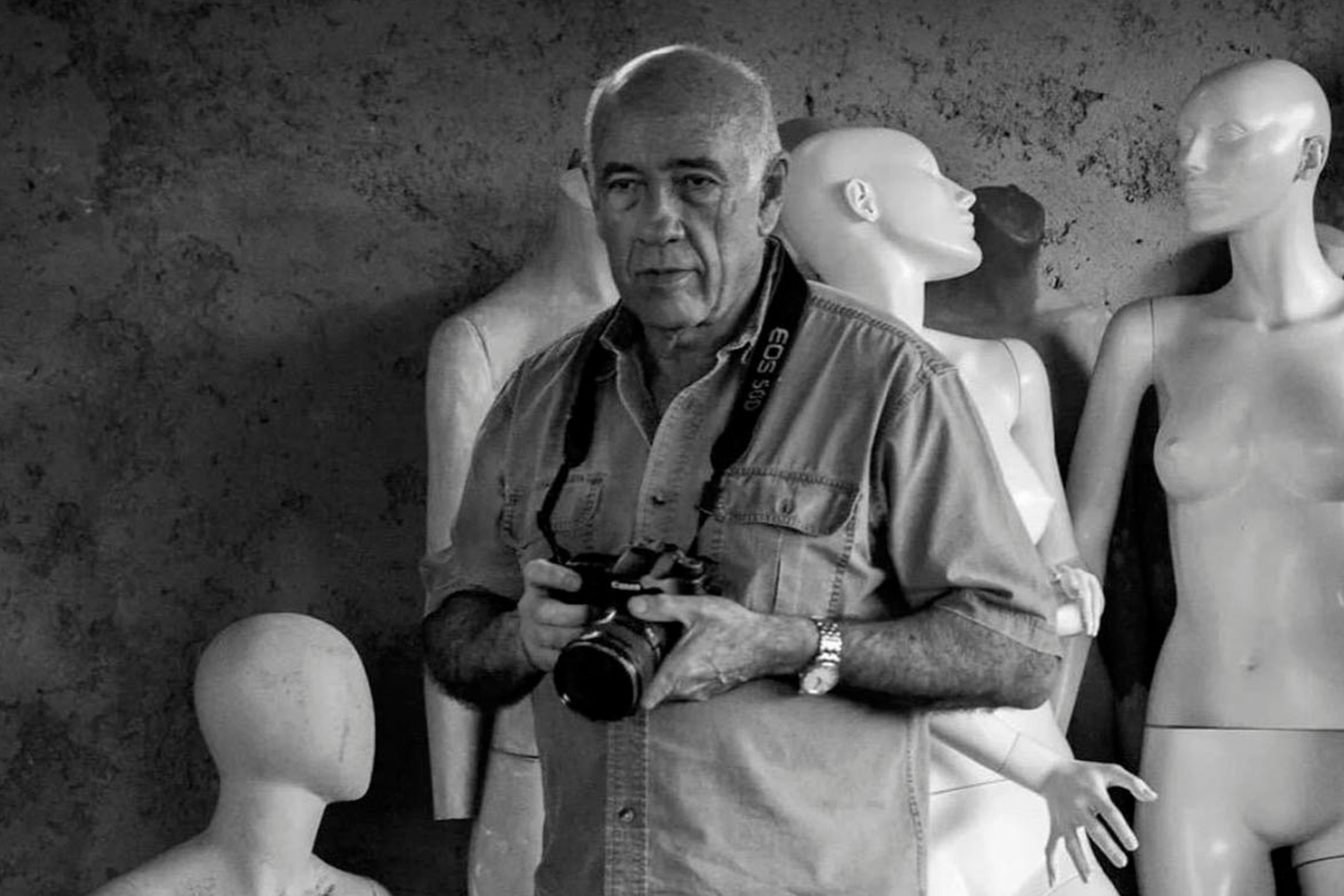 Alexis Pérez-Luna, con camisa clara y una cámara en las manos, posa entre maniquíes blancos en una foto en blanco y negro.