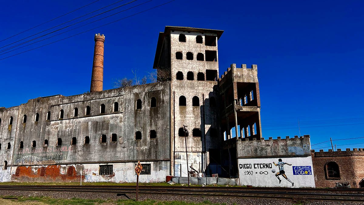 Edificio industrial abandonado con chimenea de ladrillo junto a una vía de tren, bajo un cielo azul intenso, en Cañuelas.