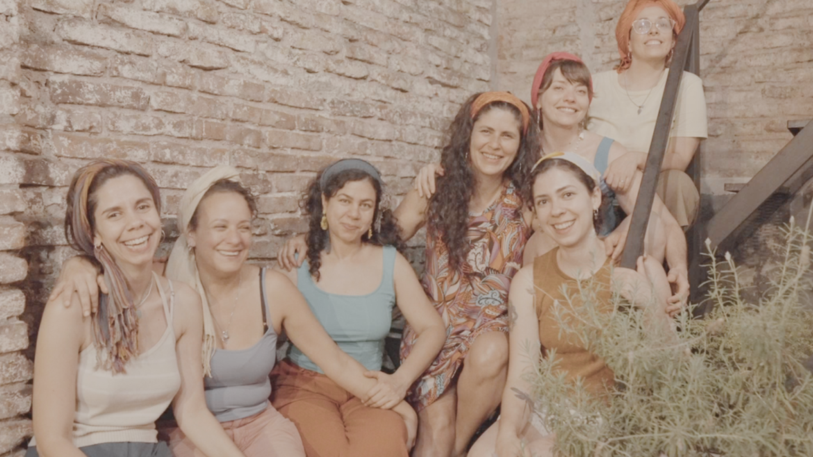 Siete mujeres con pañuelos en la cabeza posan sonrientes y abrazadas en una escalera metálica, frente a una pared de ladrillos