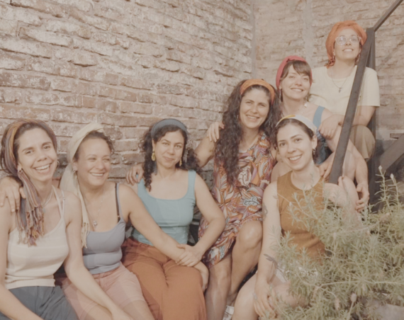 Siete mujeres con pañuelos en la cabeza posan sonrientes y abrazadas en una escalera metálica, frente a una pared de ladrillos