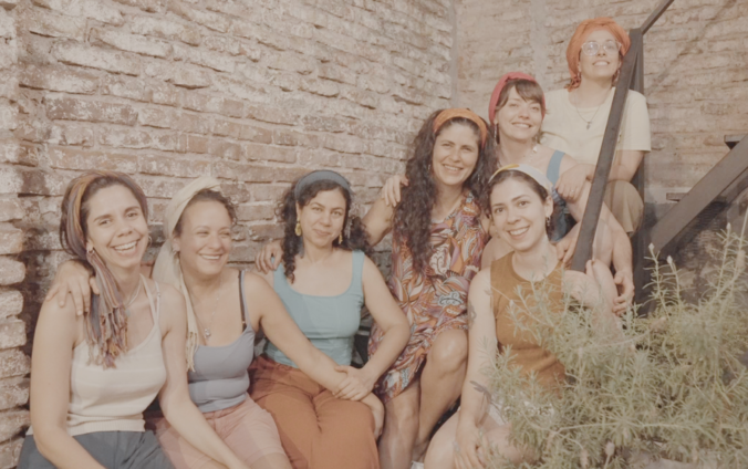 Siete mujeres con pañuelos en la cabeza posan sonrientes y abrazadas en una escalera metálica, frente a una pared de ladrillos