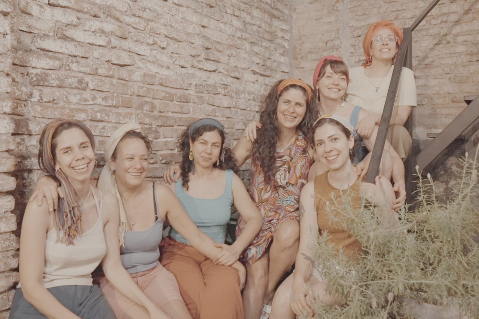 Siete mujeres con pañuelos en la cabeza posan sonrientes y abrazadas en una escalera metálica, frente a una pared de ladrillos