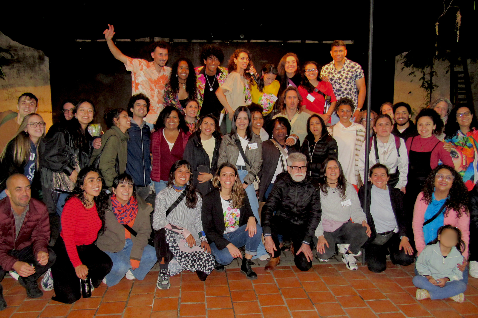 Foto grupal nocturna de artistas y participantes del Festival de Talento Migrante Latitudes en un patio con piso de baldosas