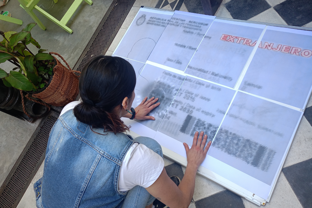 Una mujer de espaldas, agachada, apoya las manos sobre un panel que imita un documento de identidad con la palabra «Extranjero»
