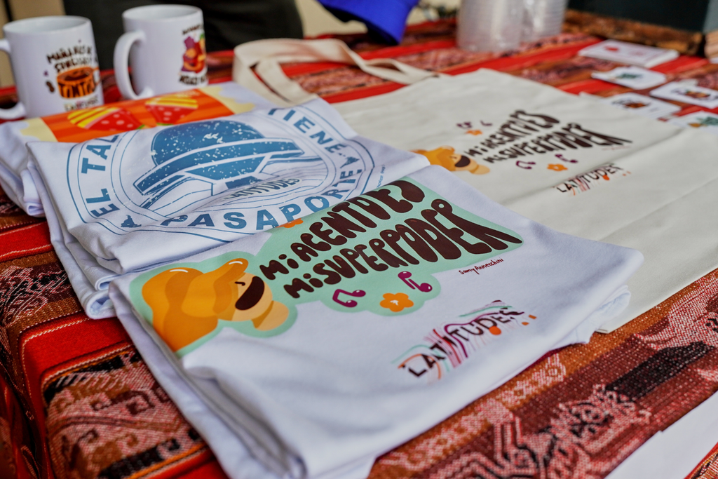 Remeras, una bolsa de tela y tazas del Festival Latitudes, con frases como «Mi acento es mi superpoder», sobre un mantel tejido