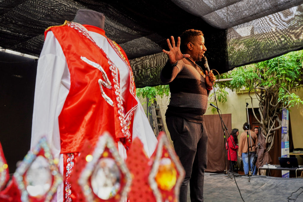Un artista habla al micrófono con una mano en alto en un escenario techado con media sombra, junto a un traje rojo con lentejuelas
