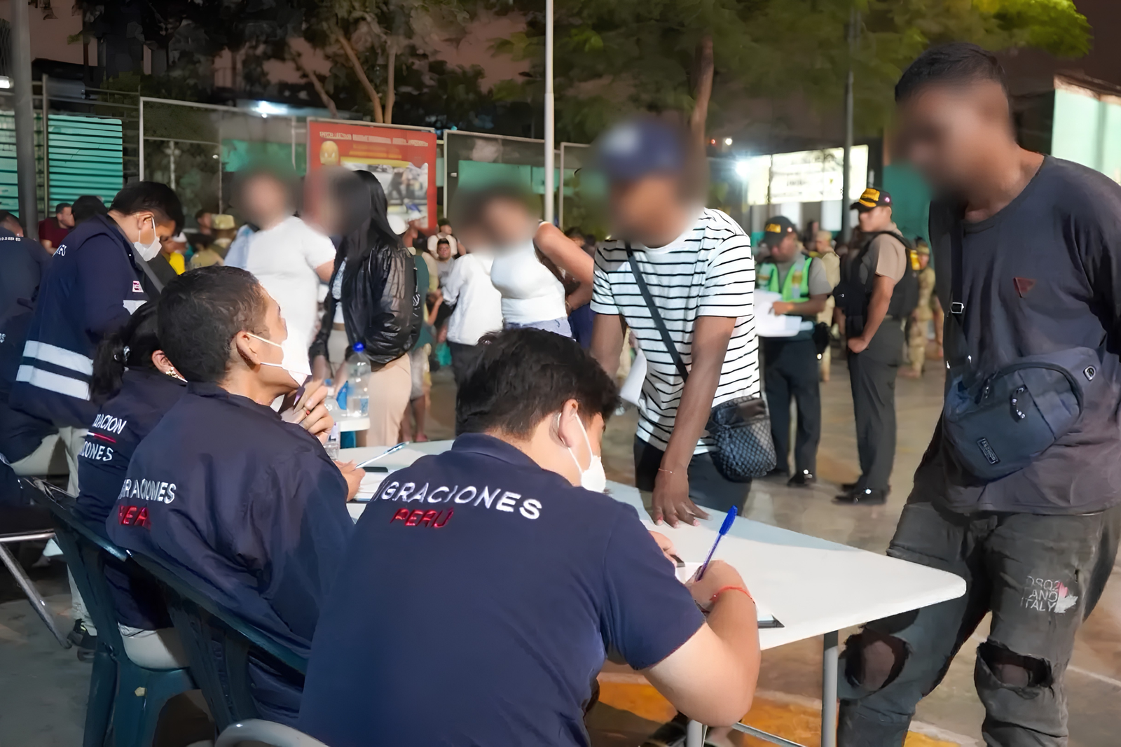 Agentes de Migraciones sentados ante una mesa registran datos de dos hombres de rostro pixelado, con policías y militares de fondo, de noche.