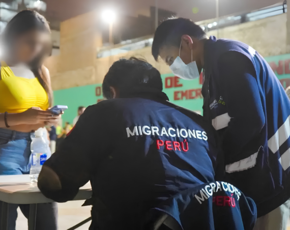 Agentes de Migraciones con chaquetas azules revisan documentos en una mesa nocturna junto a una mujer de remera amarilla con el rostro pixelado.