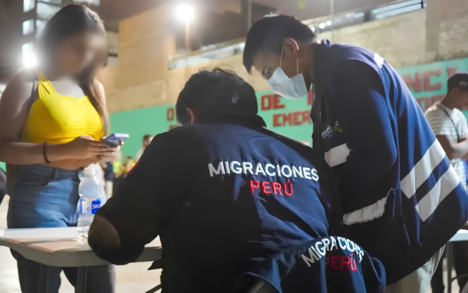 Agentes de Migraciones con chaquetas azules revisan documentos en una mesa nocturna junto a una mujer de remera amarilla con el rostro pixelado.