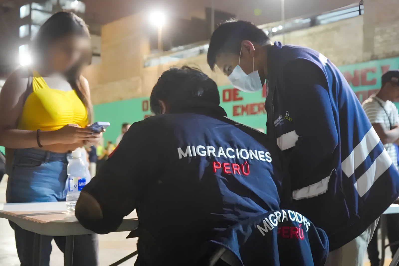 Agentes de Migraciones con chaquetas azules revisan documentos en una mesa nocturna junto a una mujer de remera amarilla con el rostro pixelado.
