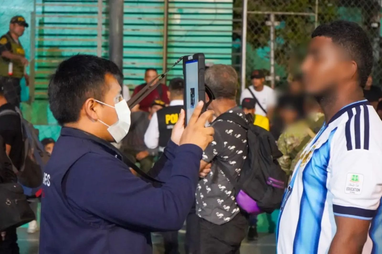 Un agente de Migraciones con mascarilla fotografía con una tableta a un hombre de rostro pixelado, mientras policías y otras personas esperan detrás.