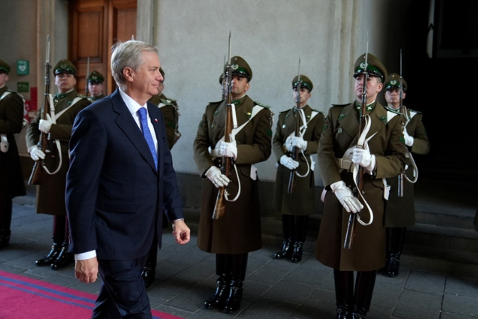 José Antonio Kast, de traje azul oscuro y corbata azul, camina por una alfombra roja frente a una guardia de honor de Carabineros en La Moneda.