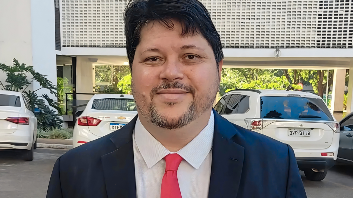 Rogério Tomaz Júnior, de traje azul oscuro y corbata roja, frente a un edificio de fachada calada con autos blancos estacionados