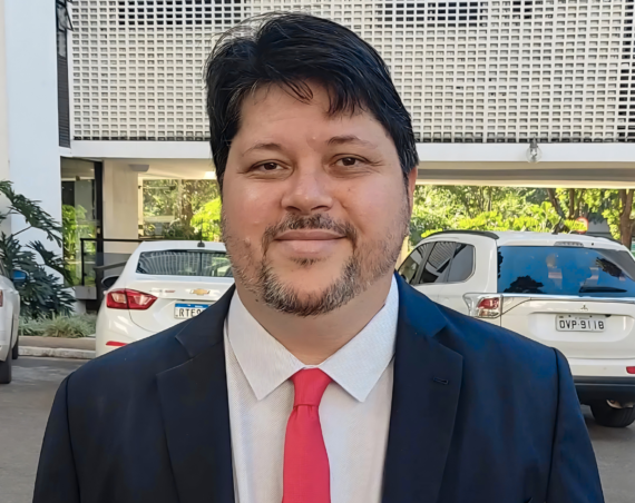 Rogério Tomaz Júnior, de traje azul oscuro y corbata roja, frente a un edificio de fachada calada con autos blancos estacionados