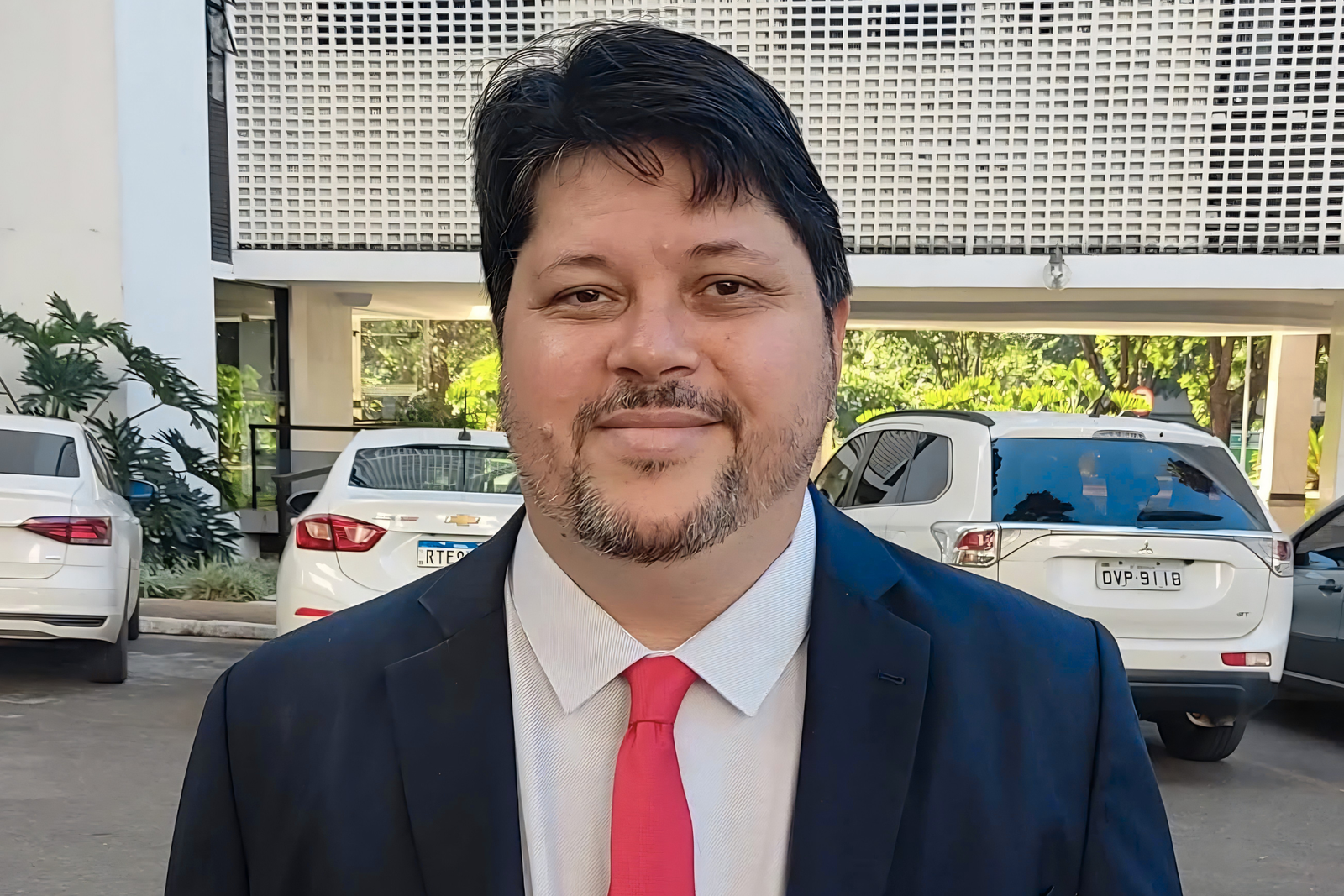 Rogério Tomaz Júnior, de traje azul oscuro y corbata roja, frente a un edificio de fachada calada con autos blancos estacionados