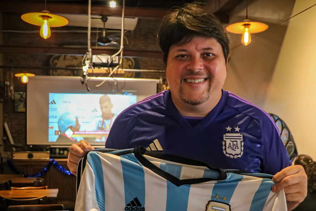 Rogério Tomaz Júnior sonríe en un bar con camiseta violeta de la AFA y sostiene una camiseta albiceleste de la selección argentina